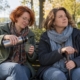 Kobieta z laską siedzi na ławce w parku, a siostra podaje jej kubek herbaty z termosu