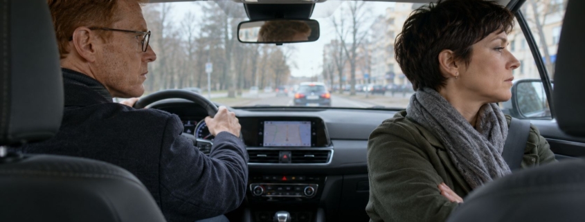 Para w samochodzie, mężczyzna prowadzi z zaciśniętymi ustami, a kobieta ze skrzyżowanymi rękami odwraca się do szyby