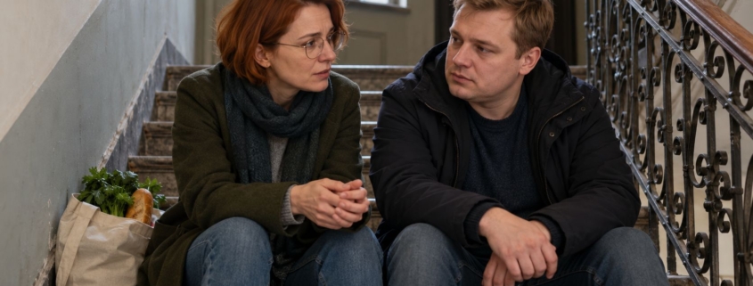 Dorosłe rodzeństwo siedzi na schodach w klatce kamienicy i rozmawia, obok stoi torba z zakupami